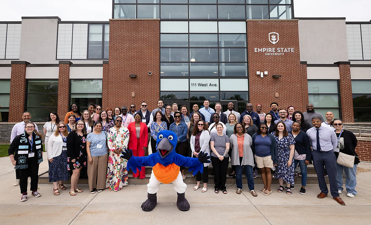a group of people stand in front of Empire State University. Blue the bluebird the mascot of Empire poses in the middle
