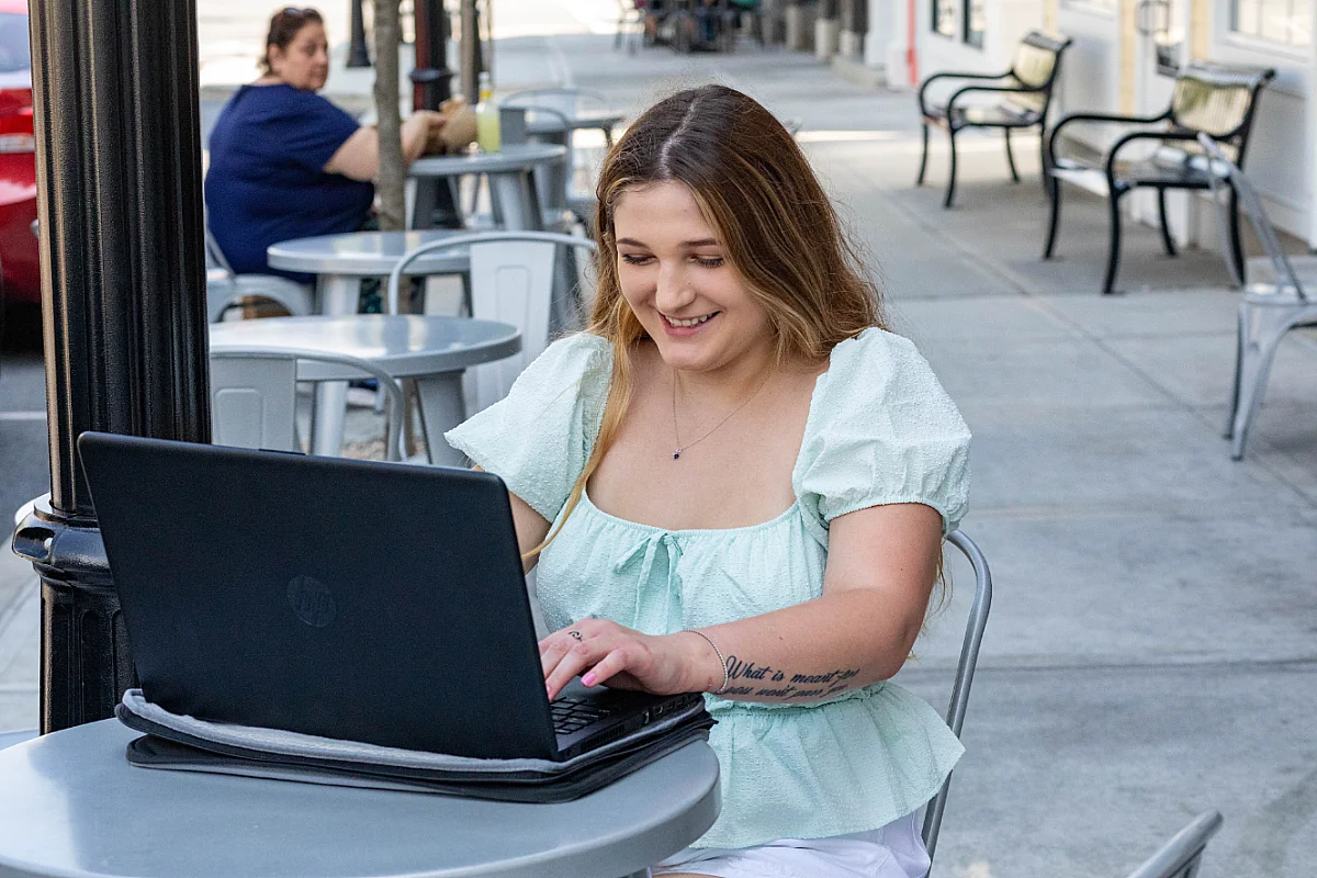 student smiles while typing on laptop at a table outside