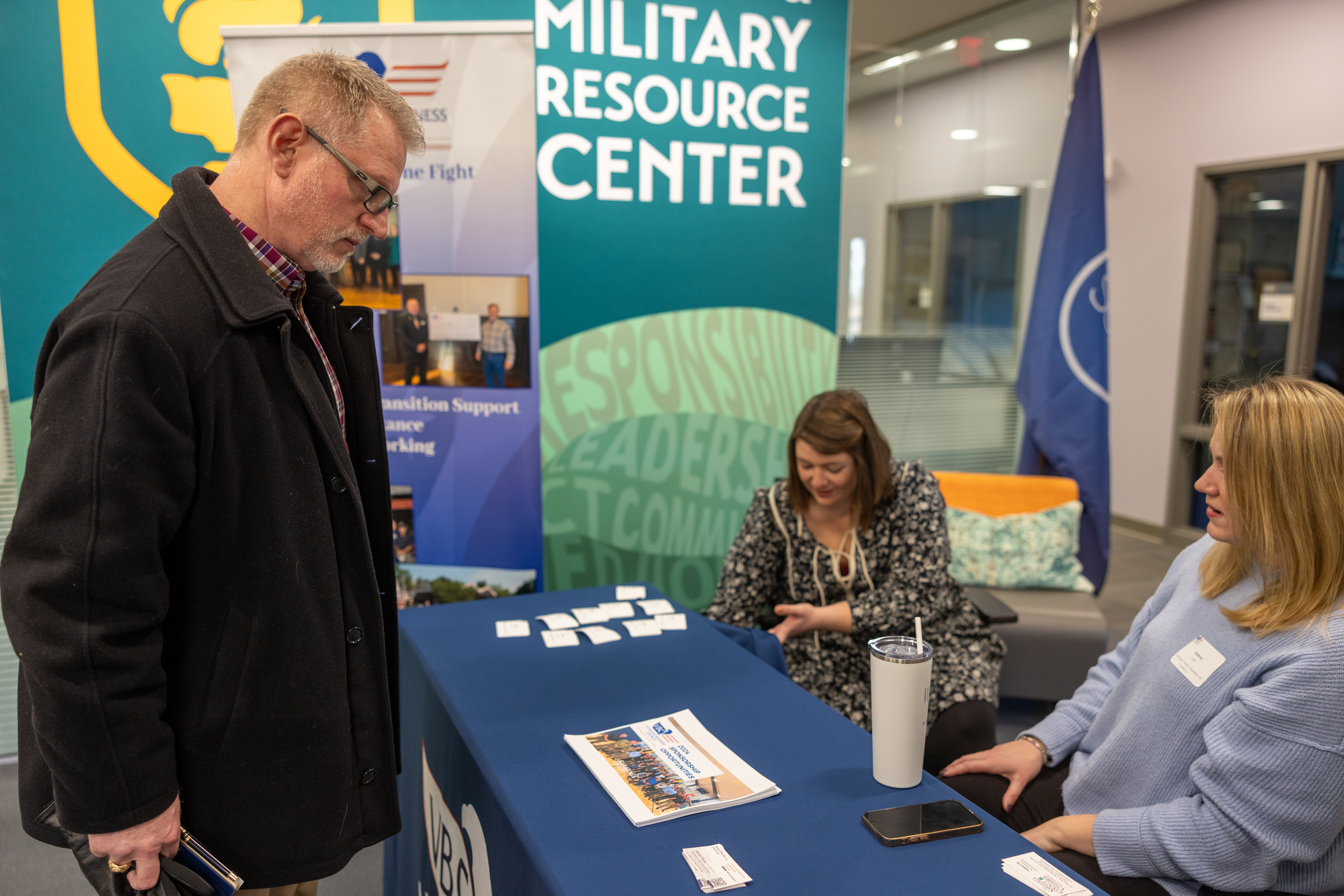 Man speaks with two female staff members at a registration table during a Veterans event.